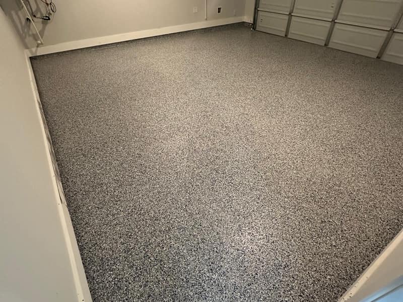 A finished two-car garage floor we coated in Katy, in an off-white and grey flake blend, looking across the bay toward the closed garage door.
